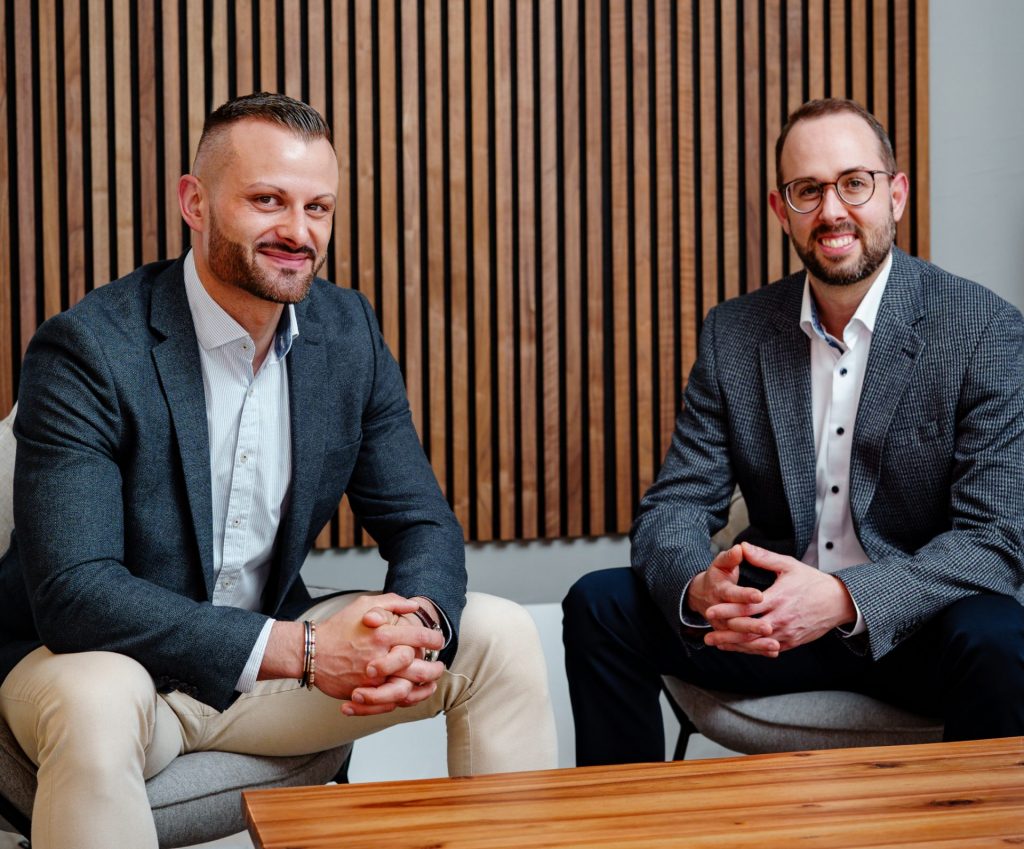 Two Exparanto team members, Nico and Markus, sitting together on armchairs at a wooden table and looking at the camera.